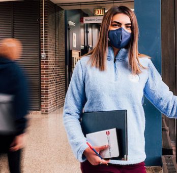 A young woman leaning against a wall with a padfolio in her hand, wearing a face mask across her nose and mouth. 