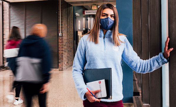 A young woman leaning against a wall with a padfolio in her hand, wearing a face mask across her nose and mouth.